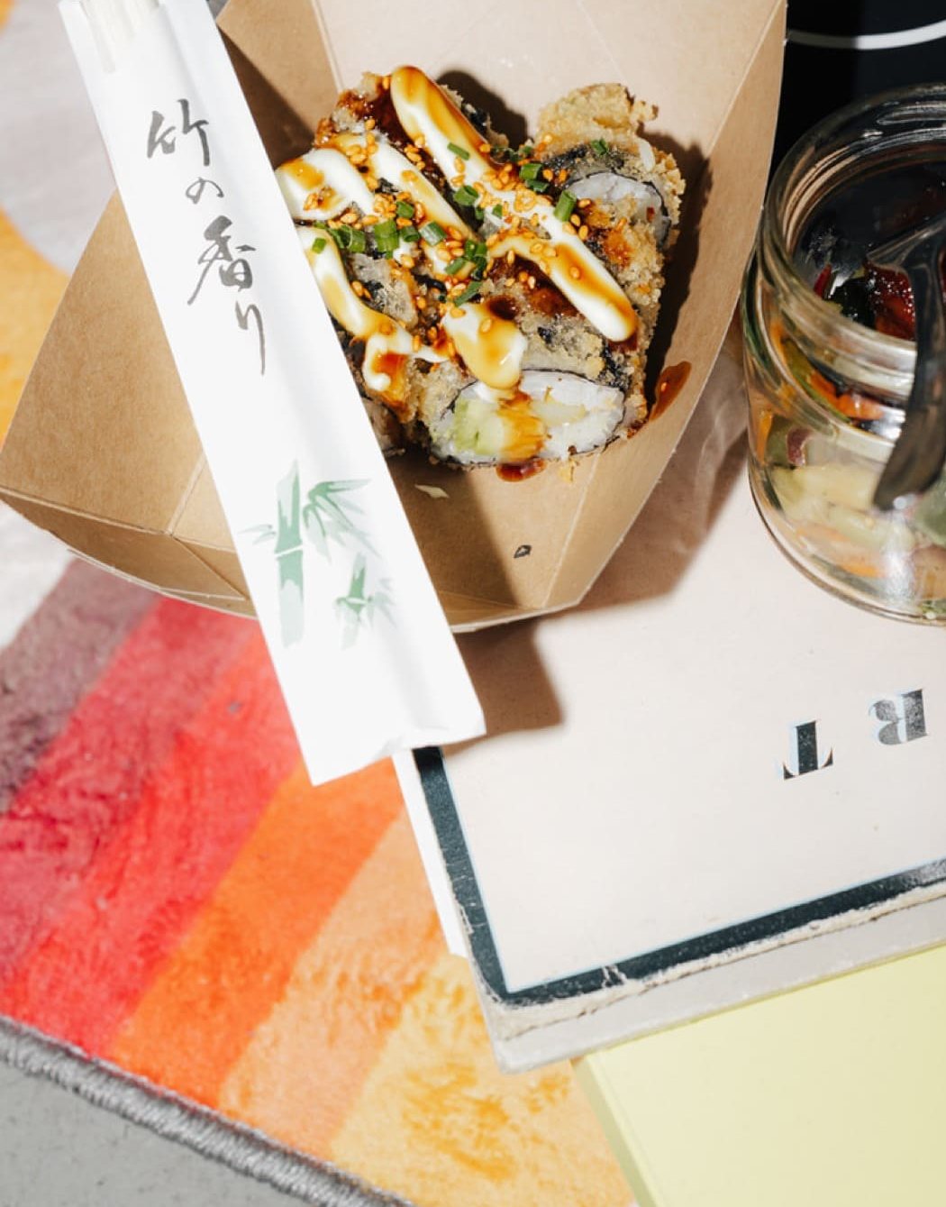 Crispy Roll mit Sesam und Sauce in der Take-away-Box aus Papier, daneben Essstäbchen und ein Glas mit Sate