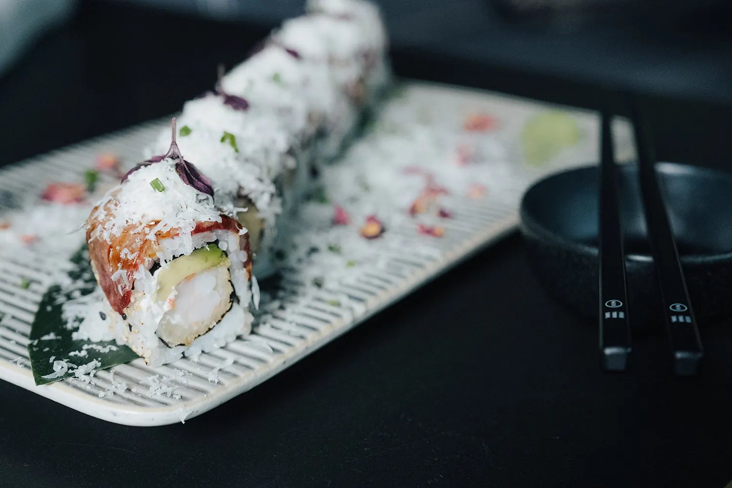 Crispy Roll mit geflämmtem Lachs und geraspeltem Meerrettich auf einer Riffelplatte, daneben schwarze Schale und Stäbchen
